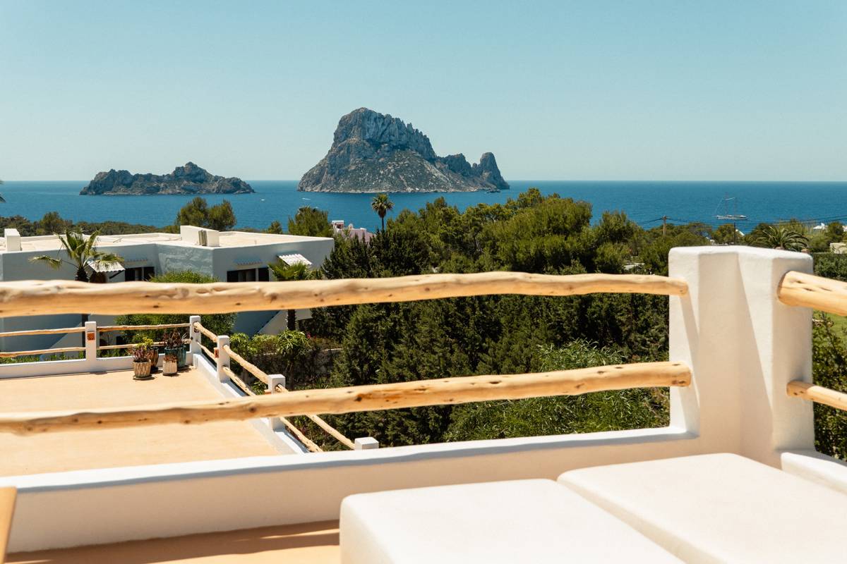 Bright terrace with wooden railings overlooking lush greenery, blue sea, and rocky islands under a clear sky.