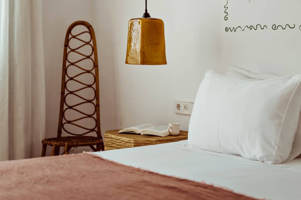 Bedroom corner with white bed, woven chair, wooden nightstand, hanging amber lamp, open book, and mug.