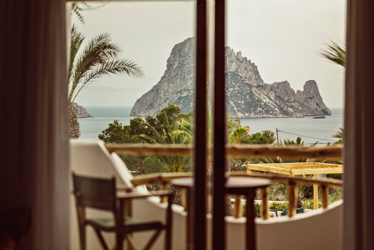 View of a rocky island and ocean through a balcony with wooden chairs, framed by palm trees.