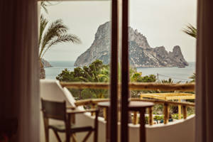 View of a rocky island and ocean through a balcony with wooden chairs, framed by palm trees.