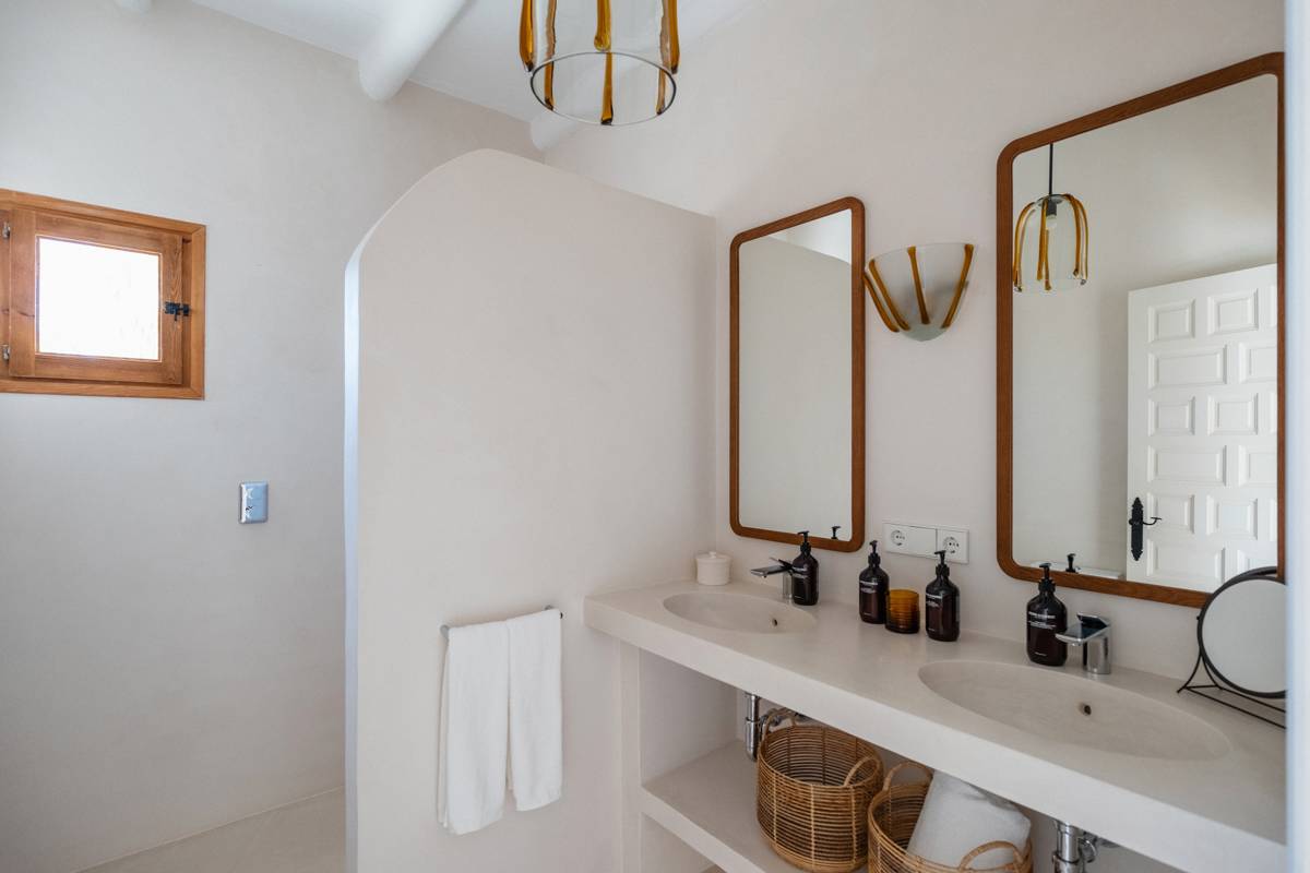 Minimalist bathroom with double sinks, two wooden-framed mirrors, wicker baskets, black soap dispensers, and a small wooden window.