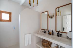Minimalist bathroom with double sinks, two wooden-framed mirrors, wicker baskets, black soap dispensers, and a small wooden window.