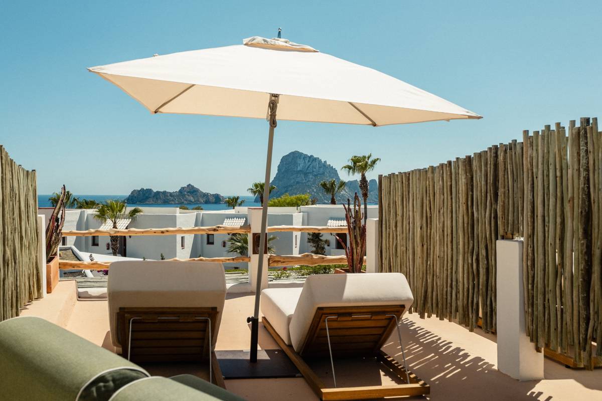 Sunny terrace with loungers under a white umbrella, overlooking blue sea, rocky islands, and white villas.
