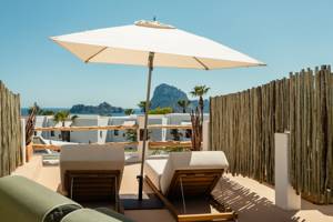 Sunny terrace with loungers under a white umbrella, overlooking blue sea, rocky islands, and white villas.