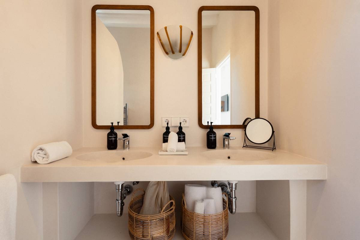 Modern bathroom with double sinks, two wooden-framed mirrors, wicker baskets, black soap dispensers, and minimal neutral-toned decor.